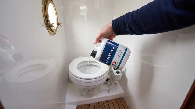 Una mano versa il disgregante Chem WC dal flacone bianco nella tazza del WC marino nel piccolo bagno di una barca a vela, con oblò in ottone e pavimento in teak