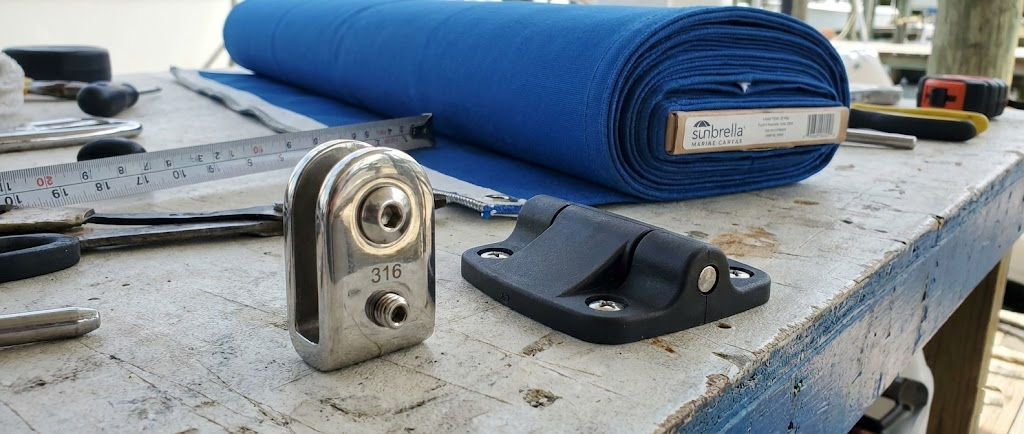 Dettaglio macro di accessori per tendalini: uno snodo in acciaio inox lucido, un attacco in nylon nero e un rotolo di tessuto nautico blu.