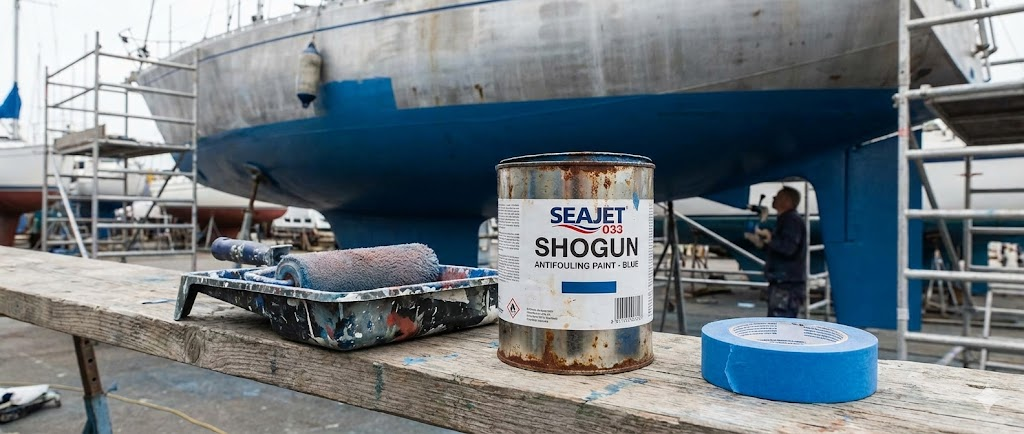 Un barattolo di antivegetativa blu con un rullo e nastro da mascheratura, sullo sfondo di una carena barca in lavorazione.