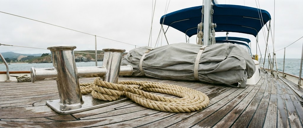 Composizione panoramica: un telo copribarca grigio piegato, un tendalino parasole aperto sullo sfondo, una bitta in acciaio inox e una cima da ormeggio.