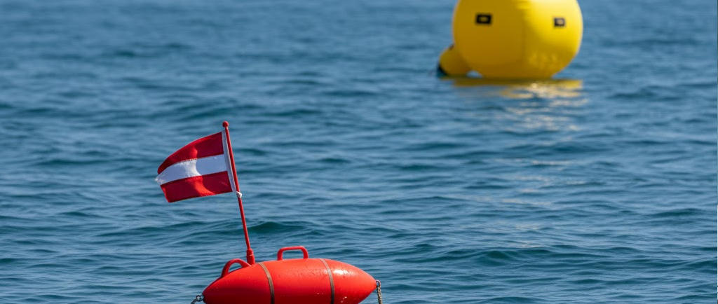Vista a pelo d'acqua: in primo piano una boa segna sub a siluro rossa con bandiera, sullo sfondo una grande boa da regata cilindrica gialla.