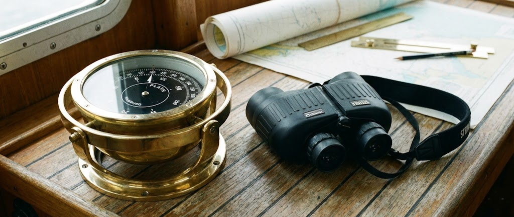 Una bussola nautica da incasso con rosa nera, affiancata da un paio di binocoli marini neri gommati e un visore notturno compatto.