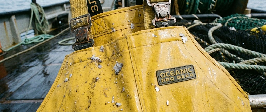 Dettaglio di una salopette in PVC giallo brillante bagnata, con fibbie robuste e rinforzi, indossata da qualcuno che tiene una cassetta di pesce.