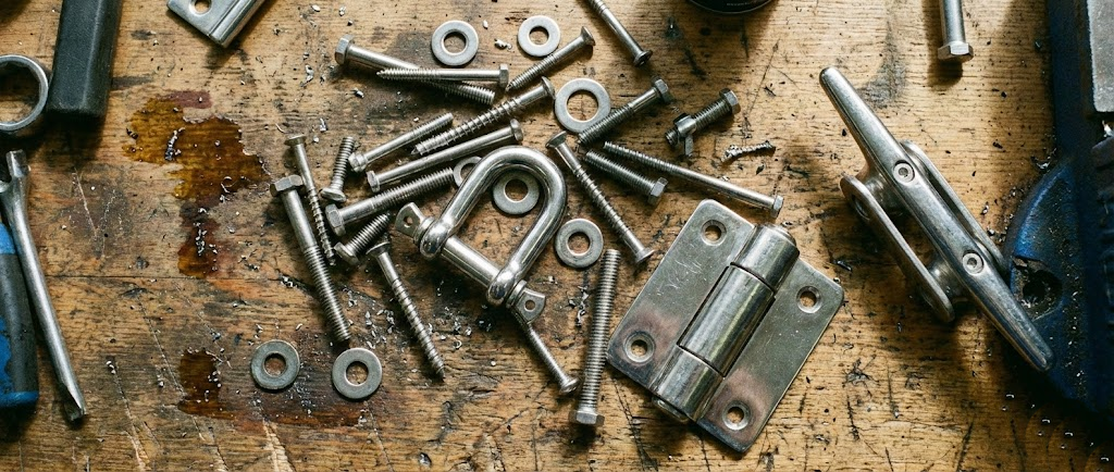 Composizione tecnica su banco da lavoro: viteria inox sparsa, un grillo lucido, una cerniera e una bitta d'ormeggio in acciaio.