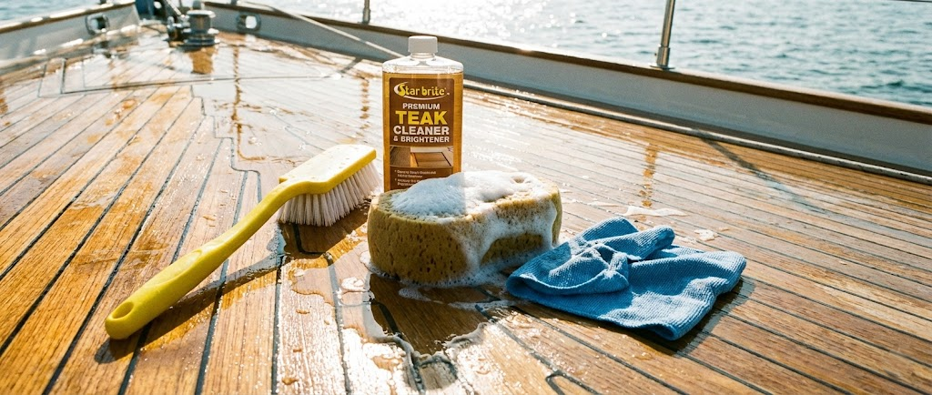 Un ponte in teak pulito con sopra un flacone di olio per teak, uno spazzolone giallo per lavaggio e una spugna insaponata.