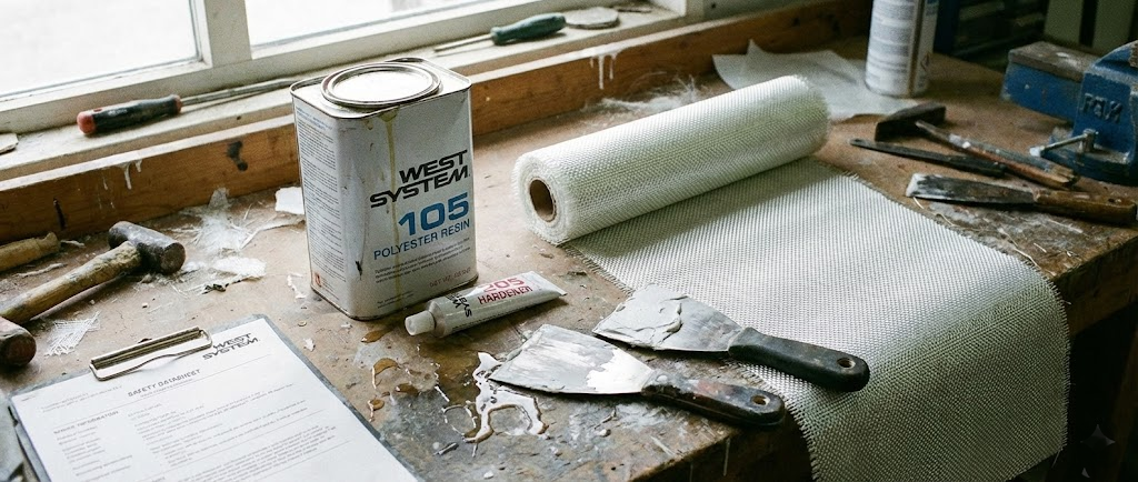 Un kit di riparazione vetroresina su un banco di lavoro: barattolo di resina, catalizzatore, tessuto in fibra di vetro e spatole per lo stucco.