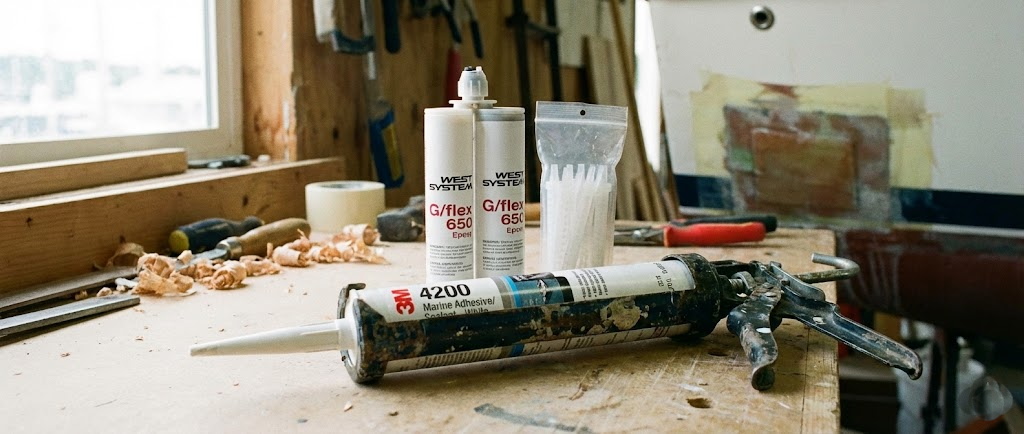 Una pistola per silicone con inserita una cartuccia di sigillante marino bianco, affiancata da tubetti di colla strutturale su un piano di lavoro.