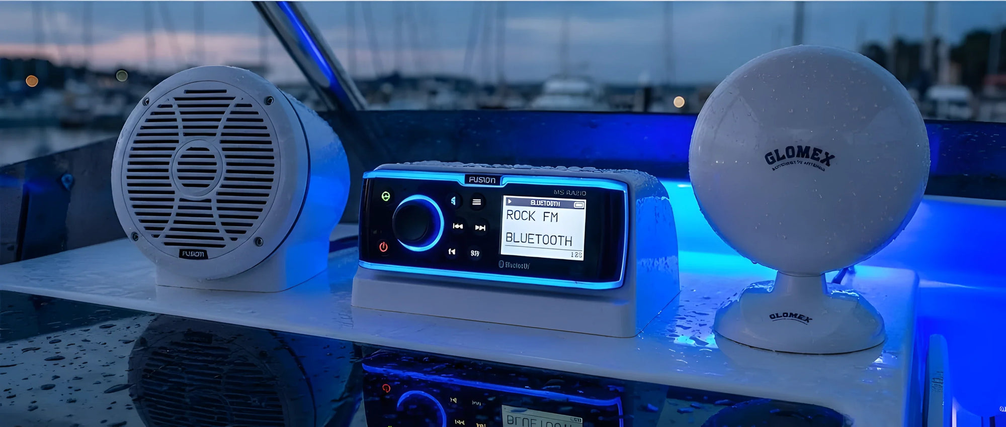 Un moderno stereo nautico con display illuminato di blu, un altoparlante bianco impermeabile e un'antenna TV a forma di disco (UFO) sullo sfondo.