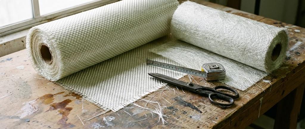 Dettaglio macro di rotoli di fibra di vetro: si vede la trama intrecciata della stuoia e la texture casuale del Mat, con forbici professionali accanto.